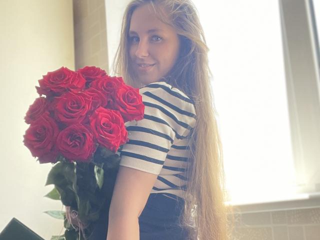 Doris mit langen blonden Haaren, einem gestreiften Hemd, die einen großen Strauß roter Rosen hält und in die Kamera lächelt. Ein helles Fenster ist im Hintergrund zu sehen. Weiches Licht..