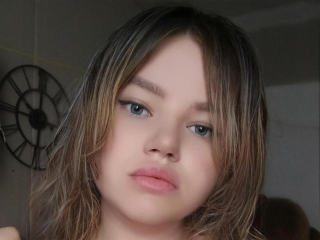 PlusSizeHt with blonde, wet-looking hair, blue eyes, and pink lips, looking slightly bored, has subtle black eyeliner, and a fair skin tone, showing a neutral expression.