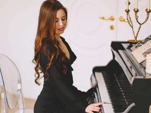 CherylFoxy with long red hair, black dress, playing a piano, her hands on the keys, her ass slightly visible, white chair and golden door in the background.