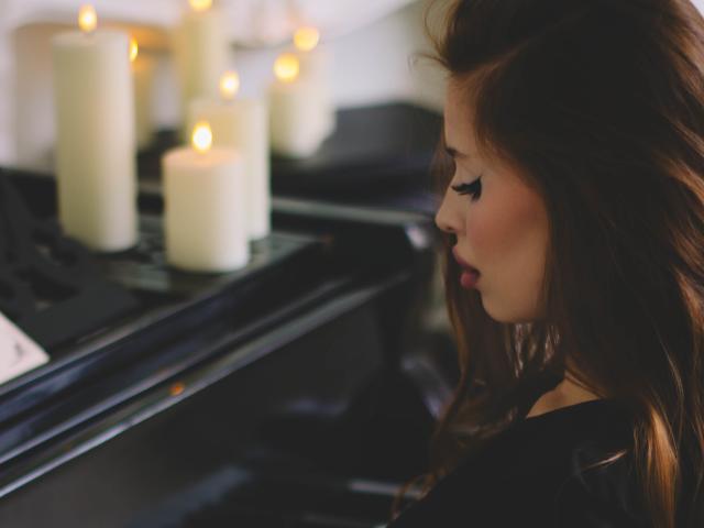 CherylFoxy with long brown hair, closed eyes, and black eyeliner, playing a black piano, surrounded by lit white candles, looking focused.