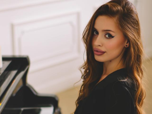 CherylFoxy with wavy brown hair, black eyeliner, and pink lipstick, looking over her shoulder at a piano, wearing a black top, showing her tits slightly.