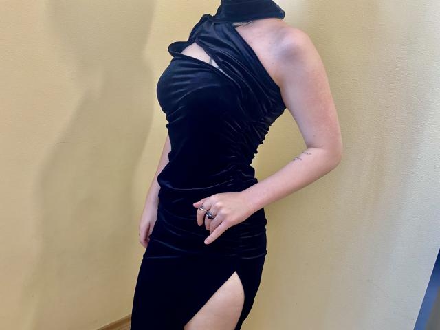 MarriAnni wearing a tight black velvet dress with a high slit, showing a tattoo on her right forearm, and a ring on her left hand.