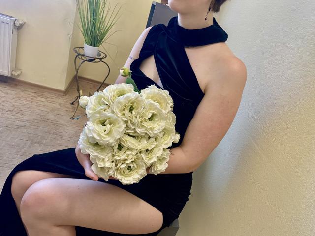 MarriAnni in a black dress holding a bouquet of white roses, sitting with her ass exposed, pale skin, short brown hair, black necklace, yellow wristwatch.
