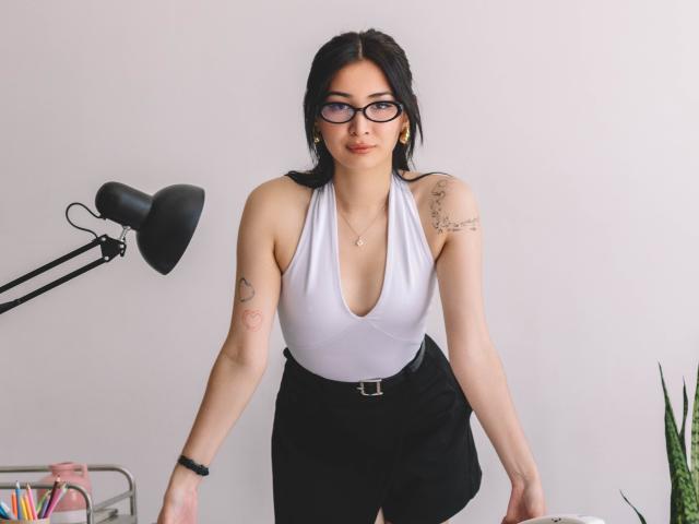 DarcyArnett with black hair, glasses, white deep V-neck top, black high-waist shorts, tattoos on arms, leaning forward, black lamp on left, green plant on right.