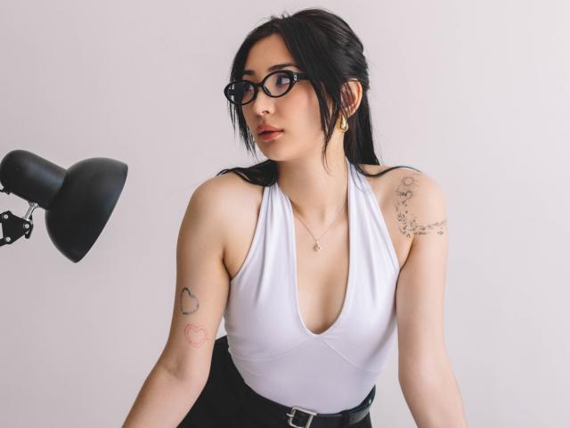 DarcyArnett has black glasses, white deep-cut top, black high-waisted pants, tattoos on arms, and black hair, leaning forward with a black lamp to her left.