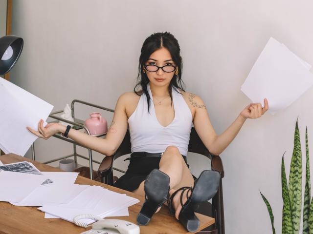 DarcyArnett, with black hair and glasses, sits in a chair, holding papers, wearing a white top, black skirt, and heels, showing tattoos, with her ass on a desk.