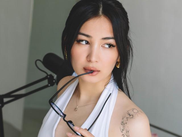 DarcyArnett with black hair, wearing a white halter top, holding glasses in her mouth, showing a tattoo on her right shoulder, and a necklace. She has a cute, sexy look.