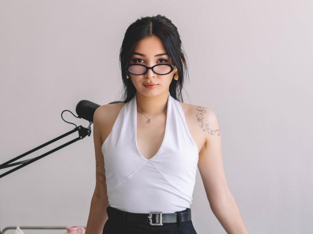 DarcyArnett with black hair, glasses, white halter top showing small tits, black pants, tattoo on right shoulder, standing, black lamp behind, light skin, gold earrings, subtle smile.