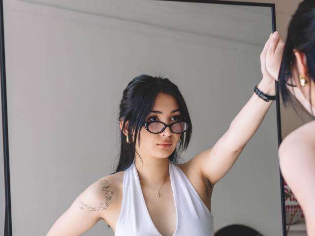 DarcyArnett with black hair, glasses, white halter top, arm tattoo, adjusting hair, wearing gold earrings, bracelet, looking in mirror, soft light, subtle smile.