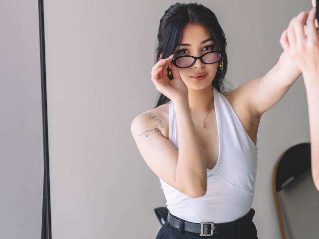 DarcyArnett with black hair, wearing a white halter top, adjusting glasses with her right hand, showing a tattoo on her left shoulder, and a black belt.