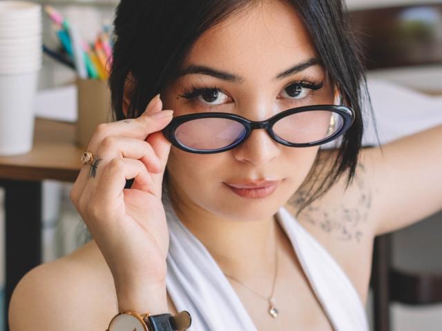 DarcyArnett with black glasses, holding them with her right hand, has tattoos on her right arm, wearing a white halter top, and a gold watch on her wrist.