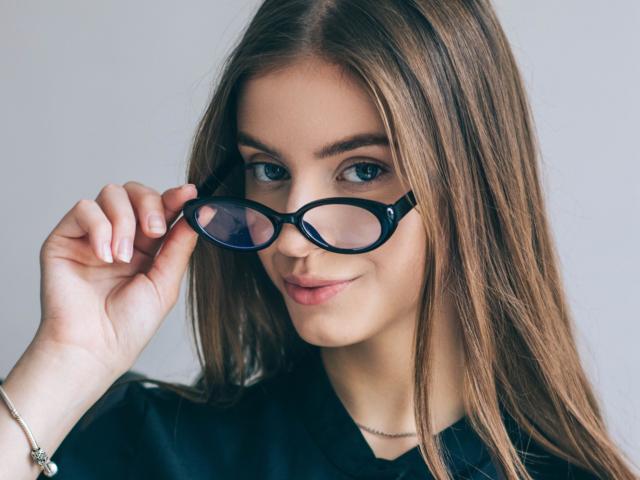EricaDiaz with long brown hair, blue eyes, and black glasses, holding them with her right hand, wearing a black shirt, and a silver bracelet, smiling subtly.