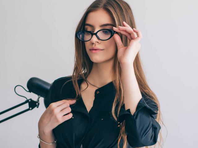 EricaDiaz with long brown hair, black glasses, and black button-up shirt, holding glasses with her right hand, smiling slightly, white background, black lamp on left.