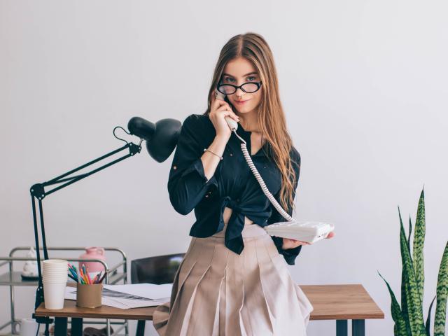 EricaDiaz with glasses, holding phone to ear, black tied shirt, beige skirt, standing by desk, lamp, papers, pen holder, green snake plant, smiling, blonde, cute, tits visible.