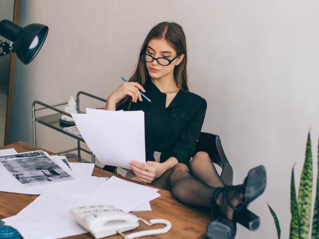 EricaDiaz with glasses, black blouse, and sheer stockings, sitting at a desk, holding a paper, writing with a blue pen, legs crossed, black heels, white phone.