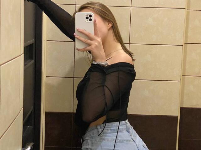JillianMel with medium tits, wearing a sheer black top and light blue jeans, taking a mirror selfie with her right arm raised, holding a white iPhone.