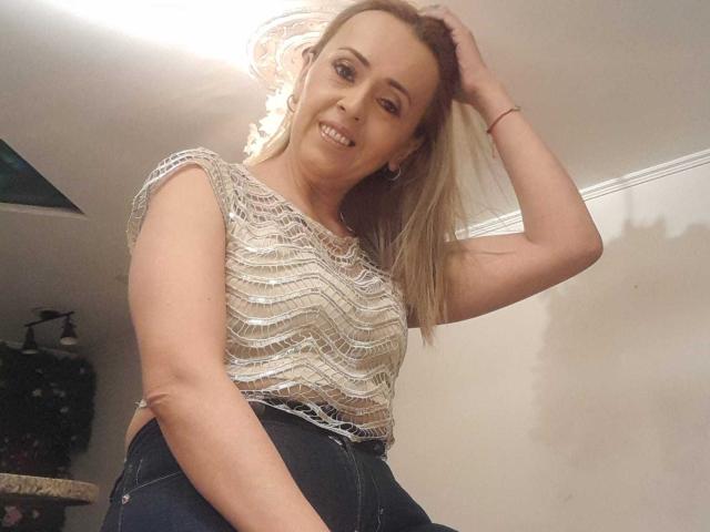 MaylinYung with blonde hair, smiling, wearing a sparkly top and black jeans, hand in hair, showing her tits, standing in a room with a ceiling light.