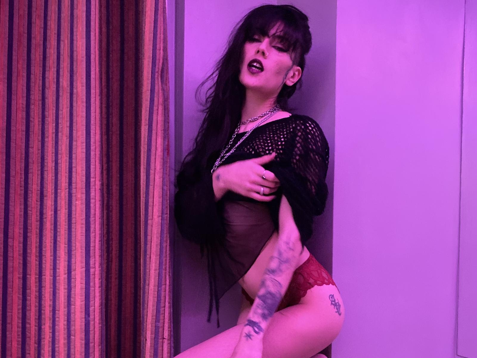 CyberDarkX with black hair, dark lipstick, black mesh top, red lace panties, tattoos on right arm and left thigh, holding chest, purple lighting, looking seductively to the camera.