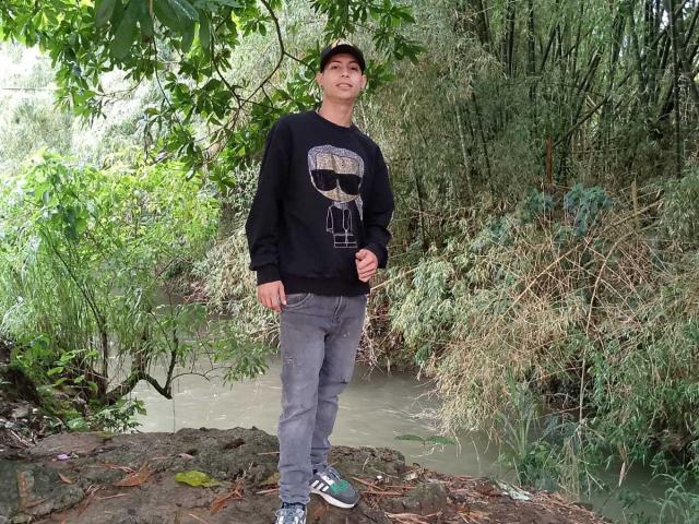 AmberAndJeff, wearing a black skull sweatshirt, gray jeans, and green/white sneakers, stands on a rock by a muddy waterway, with lush green trees in the background.