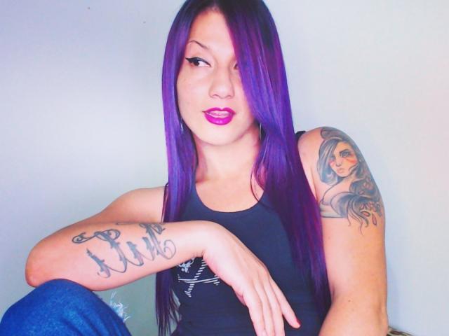 MissValentinaRamirez with purple hair, black tank top, tattoos on arm and shoulder, blue jeans, purple lipstick, winking, sitting, hand on knee, sexy, confident, attractive.