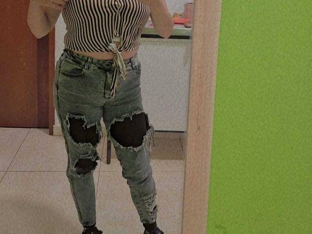 HoneyHotX indossa una maglietta a righe, annodata in vita, e jeans blu strappati con toppe nere, in piedi di fronte a uno specchio, mostrando la parte centrale del corpo e la parte inferiore..