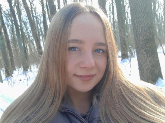 MaryannKellie has long, straight blonde hair, blue eyes, and a slight smile. She's wearing a gray jacket, and her ass is not visible. Snow and trees are in the background.