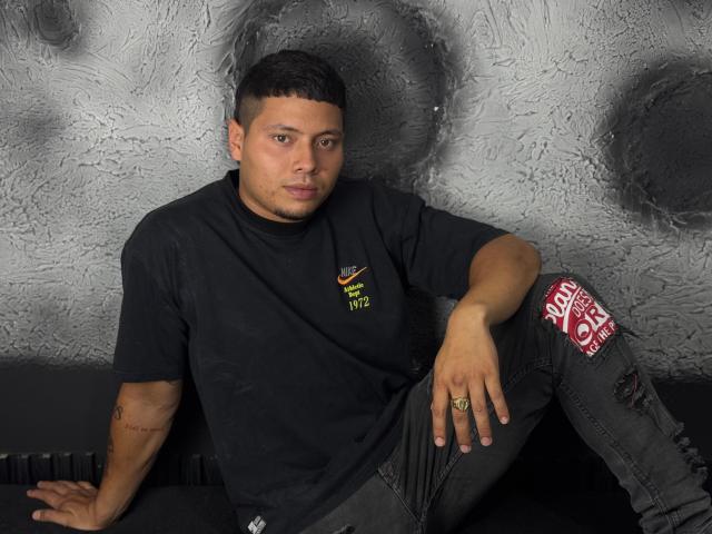 StevenClain with short black hair, wearing a black t-shirt and ripped black jeans, sitting against a textured wall, looking at the camera, with a ring on his right hand.