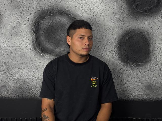 StevenClain has short black hair, a goatee, and a black t-shirt with a Nike logo and green text. His arms show tattoos, and he's looking to the side.