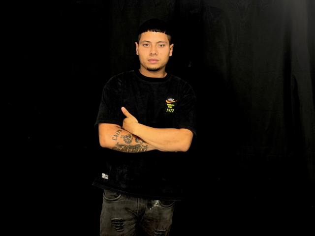 StevenClain with short black hair, wearing a black shirt and ripped jeans, standing with arms crossed, showing a tattoo on his left forearm. Black background.