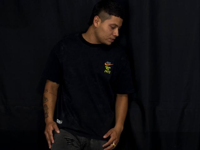 StevenClain, dark-skinned dude, short black hair, black t-shirt with small colorful logo, tattoos on right arm, looking down, black pants, hands on waist, black curtain background.