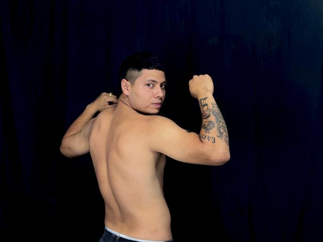 StevenClain, shirtless, flexing right arm with tattoo, black hair, looking over shoulder, wearing blue jeans, dark background, muscular back, slight smile, confident pose.