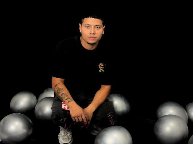 StevenClain with tattoos on his right arm, wearing a black shirt and gray sneakers, sitting among silver balloons, looking straight at the camera.