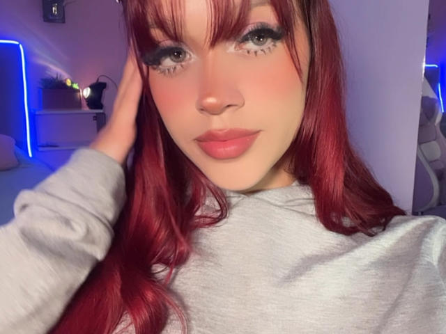 Ada Ferreira mit roten Haaren, grünen Augen und pinkem Lippenstift, trägt einen grauen Pullover, berührt ihren Kopf, hat volle Lippen und ein süßes, attraktives Gesicht. Brüste sind nicht sichtbar..