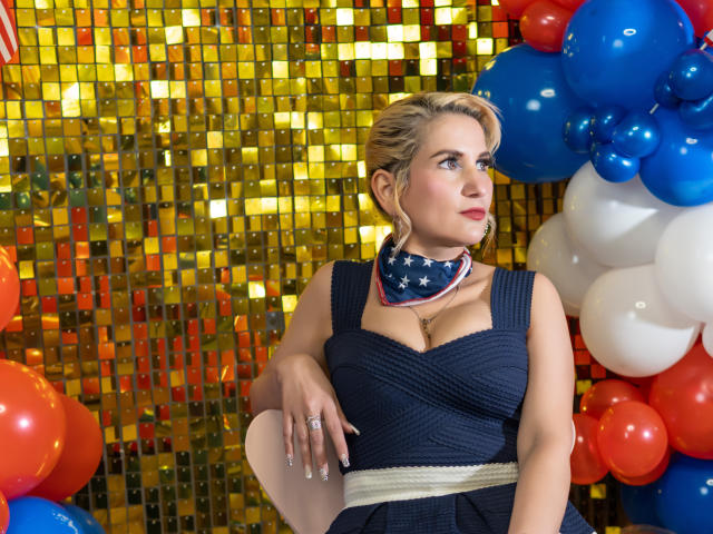 GiuliaAddams with blonde hair, blue dress, cleavage, star-patterned scarf, and red lipstick, leaning on chair, surrounded by red, blue, and white balloons. Gold sequin background.