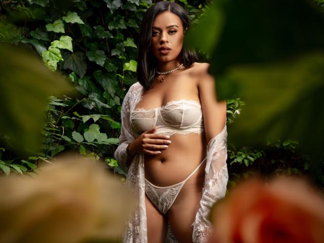 Lia Hilton, con piel morena, cabello negro, vestida con un sujetador y tanga blancos de encaje, una bata transparente, de pie en un entorno verde, con una mano sobre el vientre, un collar, mirando a la cámara, iluminación suave, sexy..
