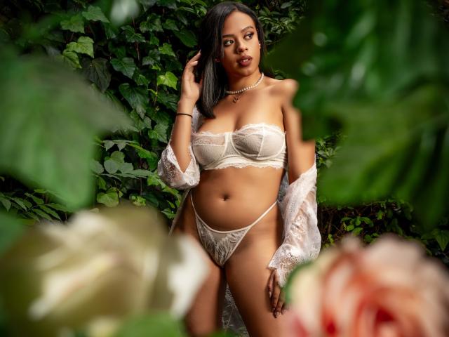 Lia Hilton, met donkere huid en lang zwart haar, draagt doorschijnende witte lingerie, een kantbeha en een string, en staat te midden van groene bladeren. Ze draagt een witte kanten badjas die van haar schouder valt, een ketting en kijkt naar de camera..