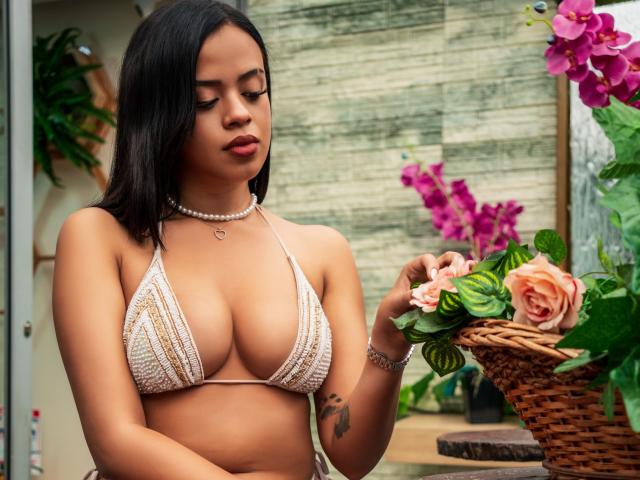 Lia Hilton, met een medium bruine huidskleur, zwart haar, draagt een witte bikini-top van gehaakt materiaal, waarbij haar borsten zichtbaar zijn, ze houdt een roze roos vast, heeft een ketting om, een tatoeage op haar rechterarm en een rieten mand..