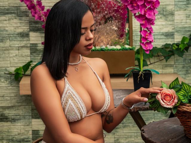 Lia Hilton, avec une peau brun clair et des cheveux noirs, portant un bikini blanc en crochet, tenant une rose rose, montrant ses seins, avec une orchidée rose en arrière-plan..