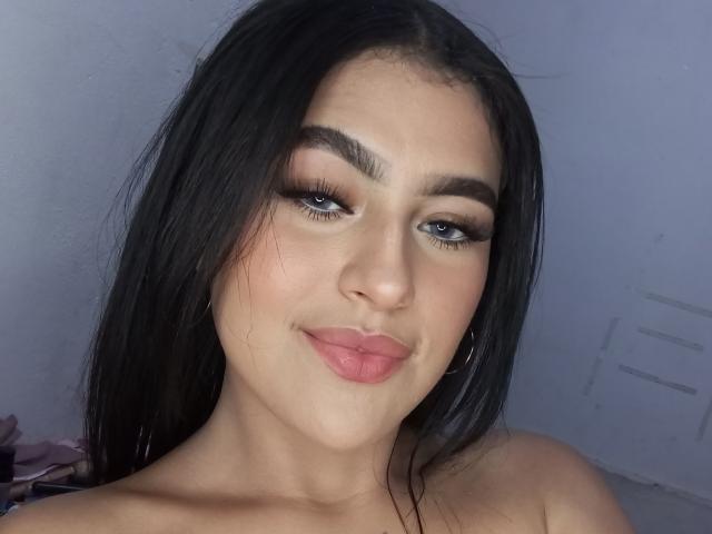 DaffneAddison has long black hair, full pink lips, and thick eyebrows with dramatic eyeliner. Her blue eyes are slightly squinted in a subtle smile. She's topless with visible collarbones.
