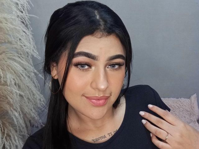 Daffne Addison, com cabelo preto, sobrancelhas grossas, cílios longos, batom rosa, blusa preta, brincos de argola, tatuagem abaixo do queixo, sorrindo, com a mão no ombro, esmalte rosa claro..