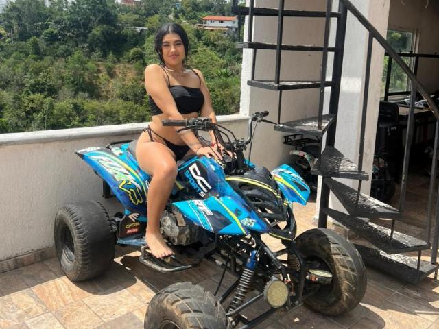 Daffne Addison, con i capelli scuri, indossa un bikini nero e siede a piedi nudi su un quadriciclo blu, verde e giallo, su un balcone con una scala in metallo..