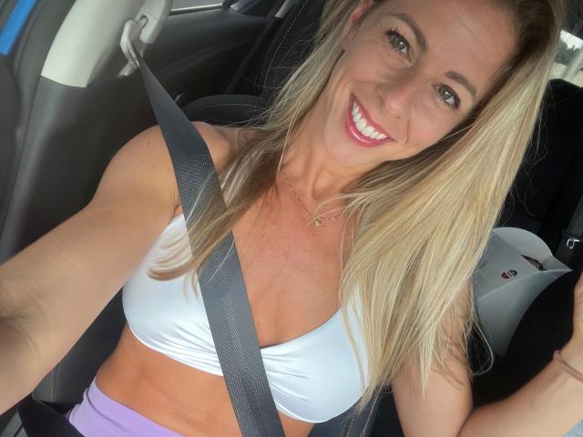 Tittney, com cabelo loiro, sorrindo, vestindo um top esportivo branco, com cinto de segurança, calças roxas, interior de carro, seios médios, corpo esguio, pele bronzeada, batom rosa e um colar de ouro..