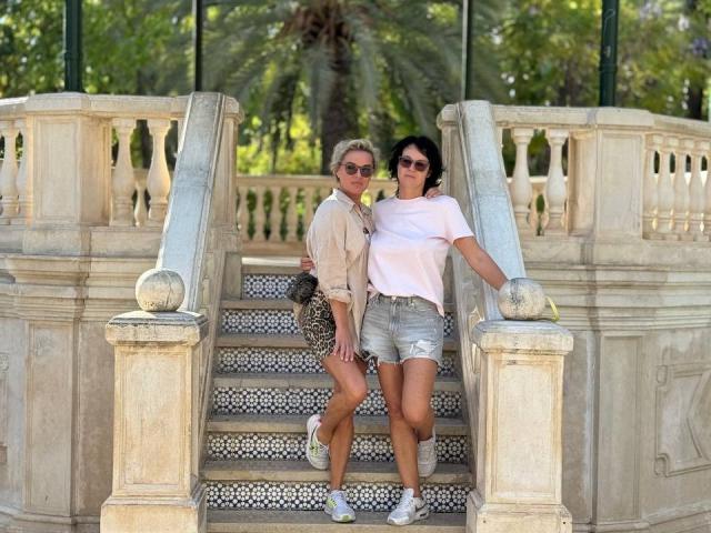 PleasureWeCouple has blonde hair, wearing sunglasses, beige shirt, leopard shorts, and white sneakers. PleasureWeCouple has dark hair, wearing white tee, denim shorts, and white sneakers.
