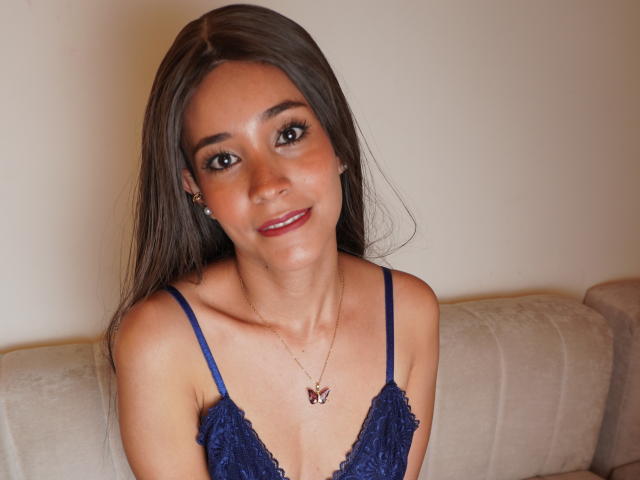 LunaMoores with long brown hair, blue lace top, small tits, red lipstick, smiling, wearing a butterfly necklace, sitting on a beige couch.