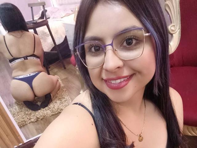 CharlotteSilvie with glasses, smiling, wearing black bra and thong, showing her ass in mirror, kneeling on rug, red chair in background, long black hair, pink lipstick.