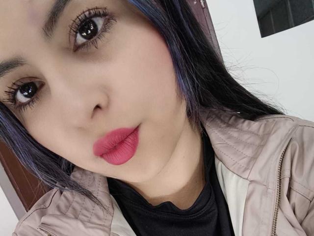 Il viso di CharlotteSilvie, con rossetto rosa, sopracciglia scure, lunghi capelli neri con riflessi viola, occhi marroni e una giacca rosa, che guarda verso la fotocamera. Il seno non è visibile..