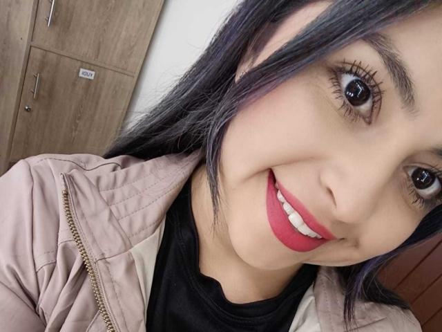 Il viso sorridente di CharlotteSilvie, con rossetto rosa, ciglia nere e capelli neri lisci, indossa una giacca beige sopra una camicia nera, mostrando il suo carino sorriso rotondo con i denti bianchi..