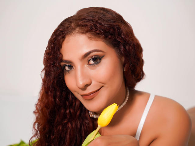 Mia Bianchi, com cabelos ruivos cacheados, segurando uma tulipa amarela, vestindo uma blusa branca com alças e um colar de pérolas, sorrindo para a câmera, exibindo seu rosto bonito e atraente..