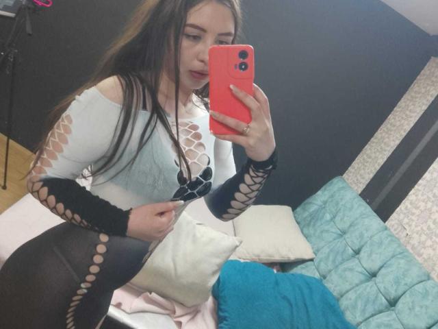 MaddieMooree with long brown hair, wearing a white lace-up top, black fishnet sleeves, and black tights, holding a red phone, taking a mirror selfie, showing her tits.