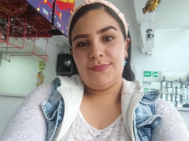 SalmaLopez, a chubby Latina woman with brown eyes, wearing a pink headband, white lace top, and blue denim vest, smiling at the camera, showing pink lipstick.